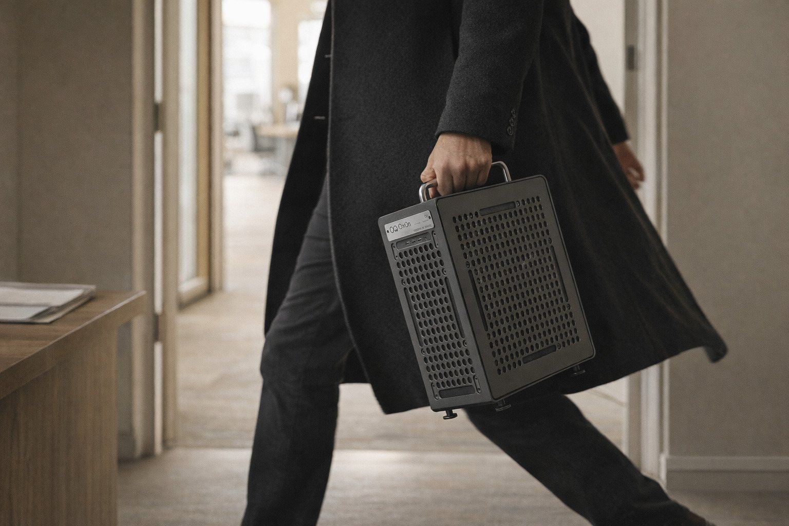A person in a dark coat walking through an office doorway, carrying the Ox1 by its handle.